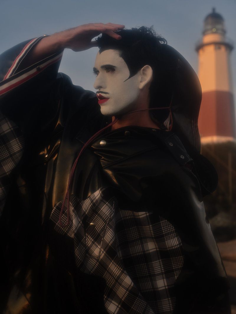 Adult male model with white face makeup, wearing black leather jacket and plaid shirt, poses near a red and white lighthouse.