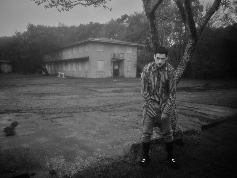 Monochrome portrait of a man with makeup and striped shirt standing in front of a building, with trees and a wet ground.