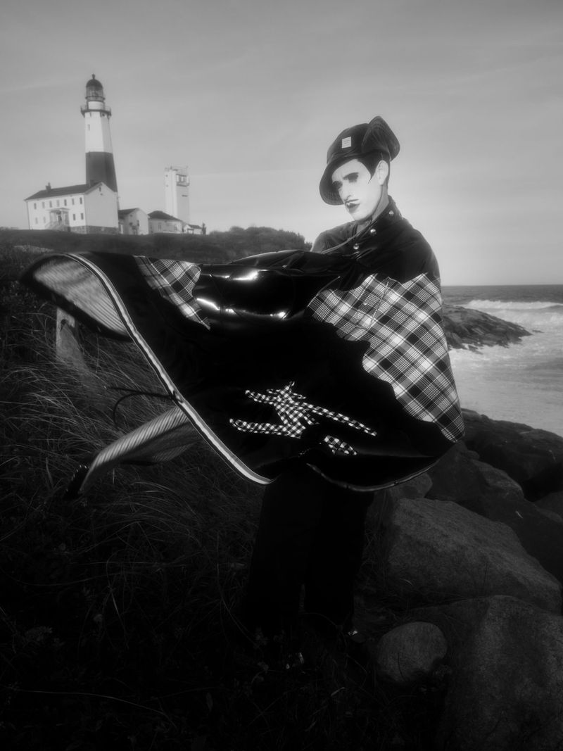 Black and white portrait of a person in makeup and theatrical clothing, holding a blanket on rocks near the ocean with a lighthouse in the background.