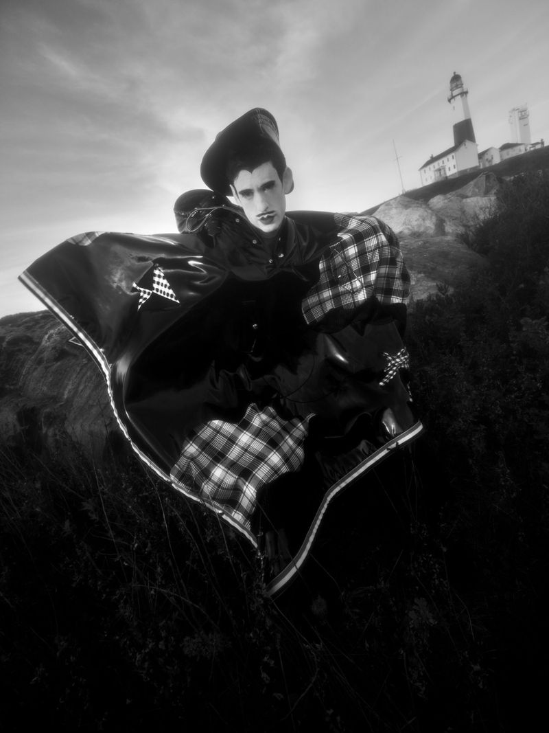 Monochrome portrait of a person in distinctive clothing and hat, posed outside with a lighthouse in the background. Cloudy sky.