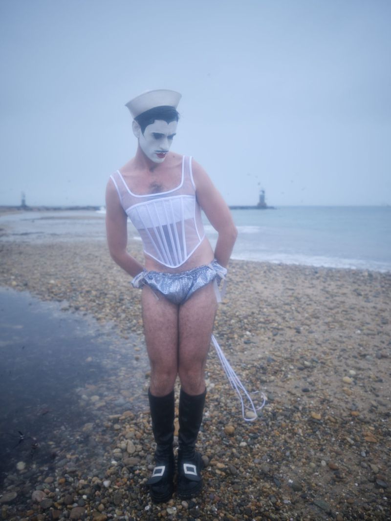 A person in white face paint, wearing a sailor hat, white corset, and silver underwear, stands on a rocky beach near the water.