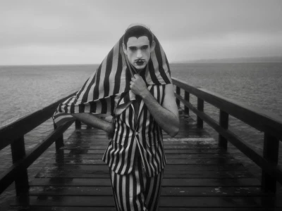 Man in black and white striped suit with theatrical makeup posing on a wooden pier extending into the water