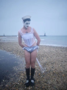 Model in theatrical white makeup, a sailor cap, sheer boned corset, and ruffled shorts on an overcast pebble beach.
