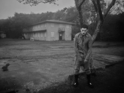 Monochrome photograph of a styled male model in a striped shirt and dark boots standing near a tree in front of an abandoned building.