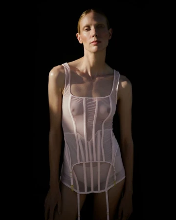 A woman with fair skin and blonde hair poses in lingerie against a black backdrop, highlighted by a spotlight that accentuates her figure.