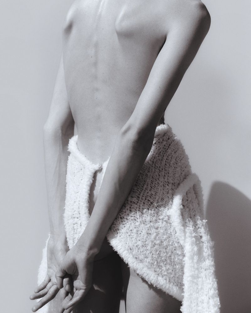 Artistic nude back shot of a person with arms crossed with a white, woven cardigan draped over the lower body against a neutral background.