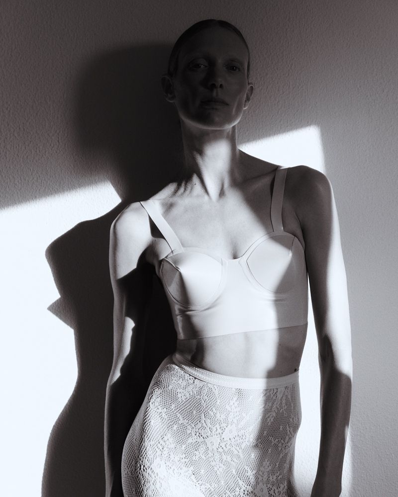 Monochrome portrait of a person with close-cropped hair wearing a bra top and lace skirt, posing against a light wall with shadow.