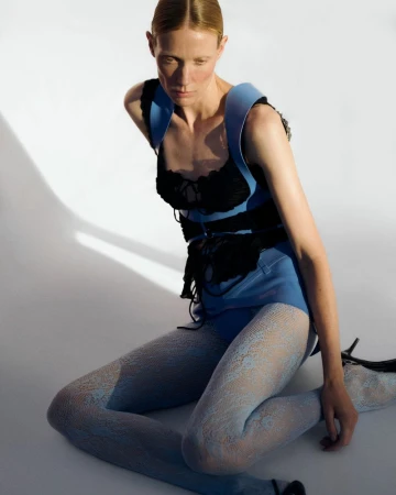 Annely Bouma seated on a white floor wearing a blue structured top, black lace bodice, and patterned blue tights.