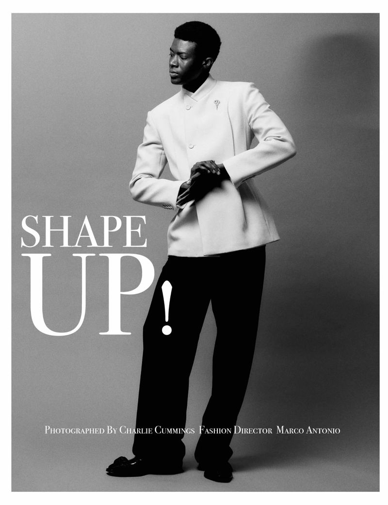 Full-length portrait of a Black male fashion model in a white suit jacket with black pants in a studio setting, with a grey background.