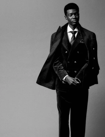 Portrait of an adult Black male in a suit with a tie and coat standing indoors in front of a grey backdrop in black and white.