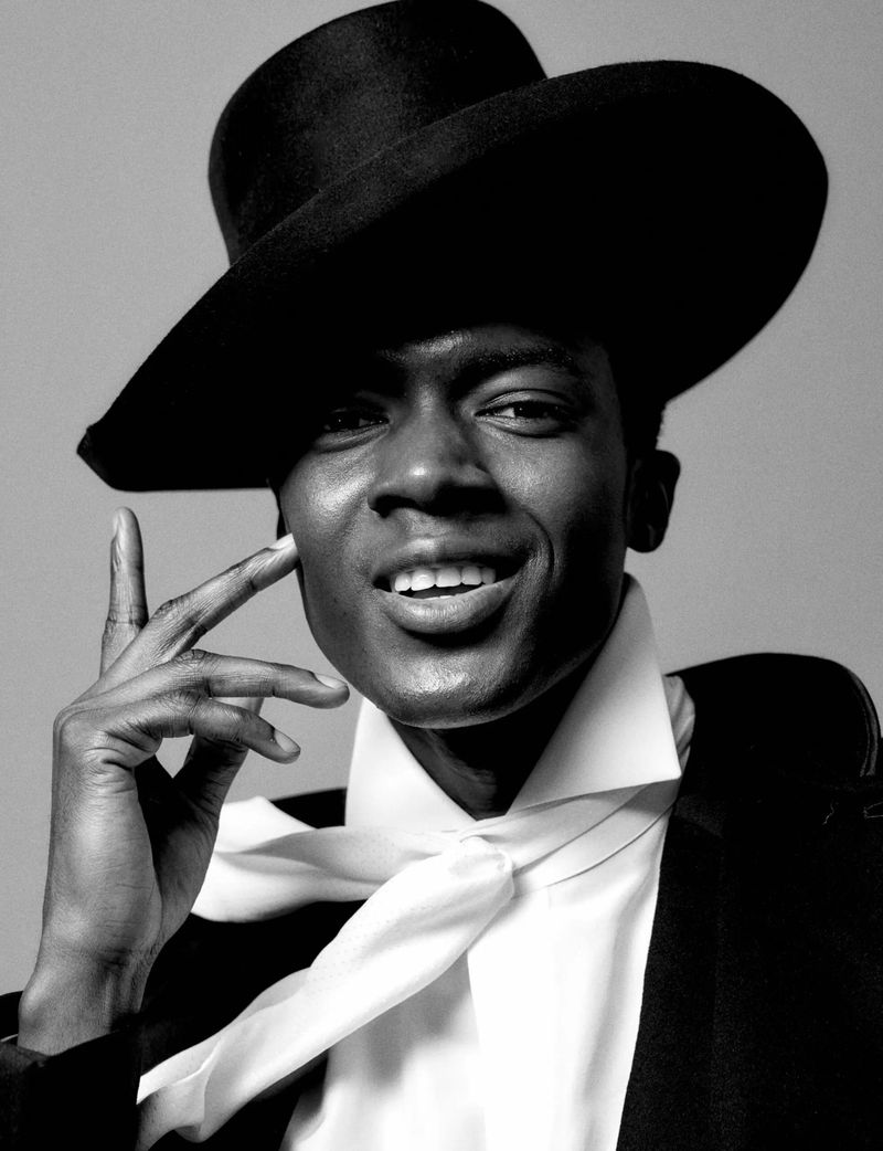 Black and white portrait of an adult male wearing a hat, suit jacket, and tie, holding fingers to his face with a slight smile.