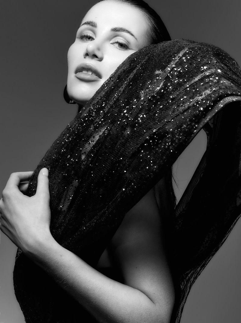 Black and white portrait of a woman wearing a shiny, textured shawl, looking upwards. The image has high contrast and highlights the model's features.