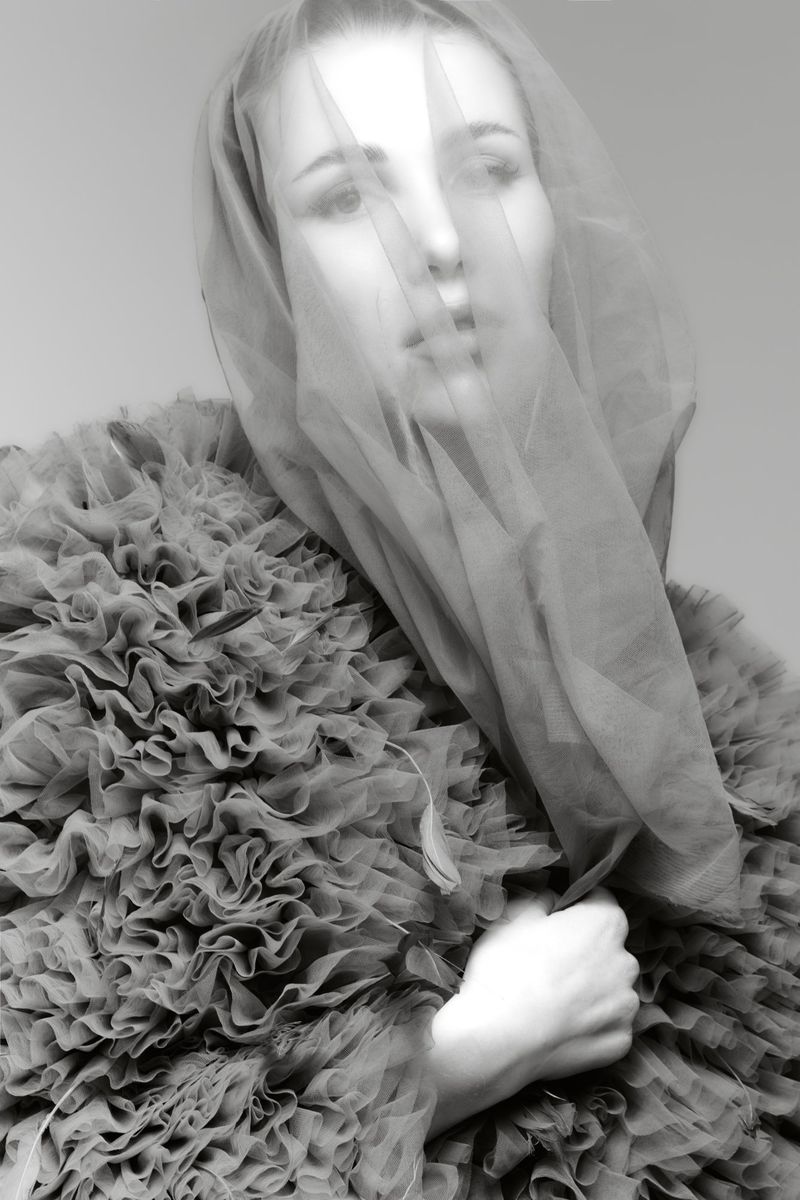 Monochrome portrait of a fair-skinned adult female model wearing a ruffled jacket, with a sheer veil covering her face and shoulders.