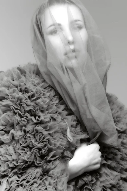 Monochrome portrait of a fair-skinned adult female model wearing a ruffled jacket, with a sheer veil covering her face and shoulders.