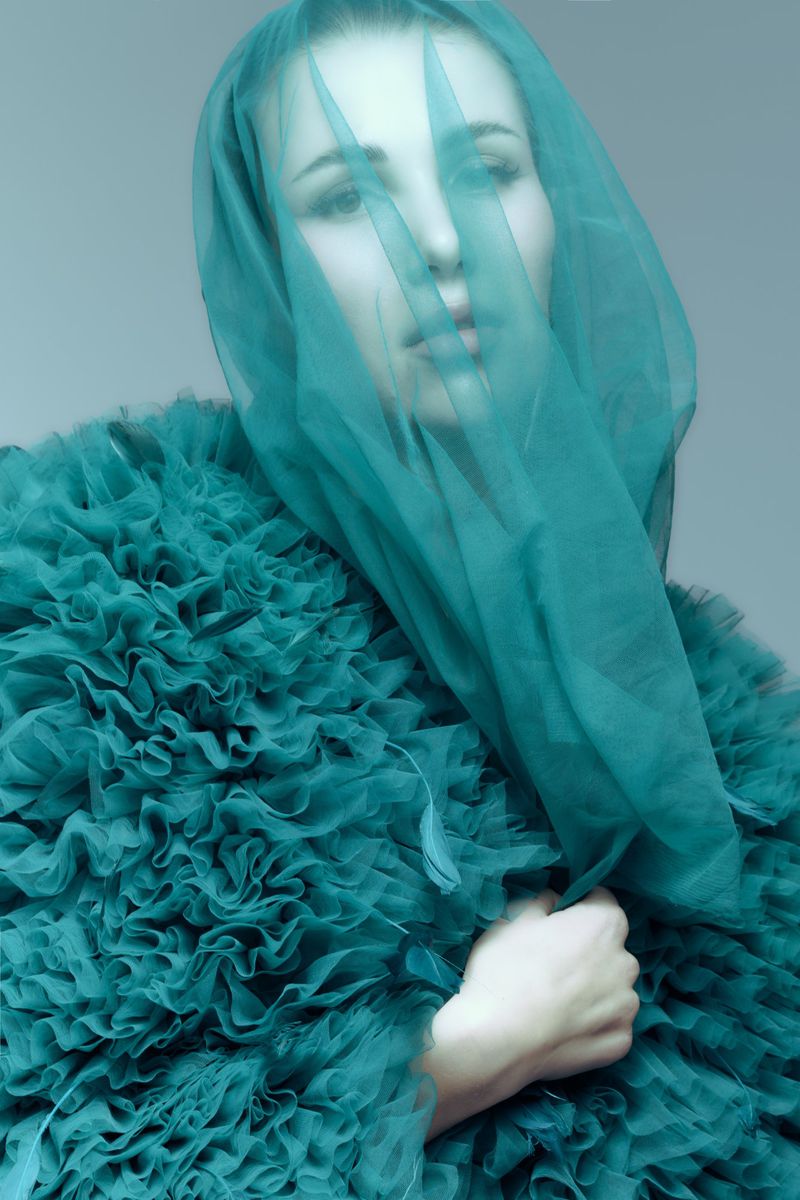 Portrait of a woman in a textured, teal dress and matching veil covering her face, against a light grey studio backdrop.