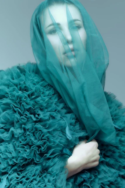 Portrait of a woman in a textured, teal dress and matching veil covering her face, against a light grey studio backdrop.