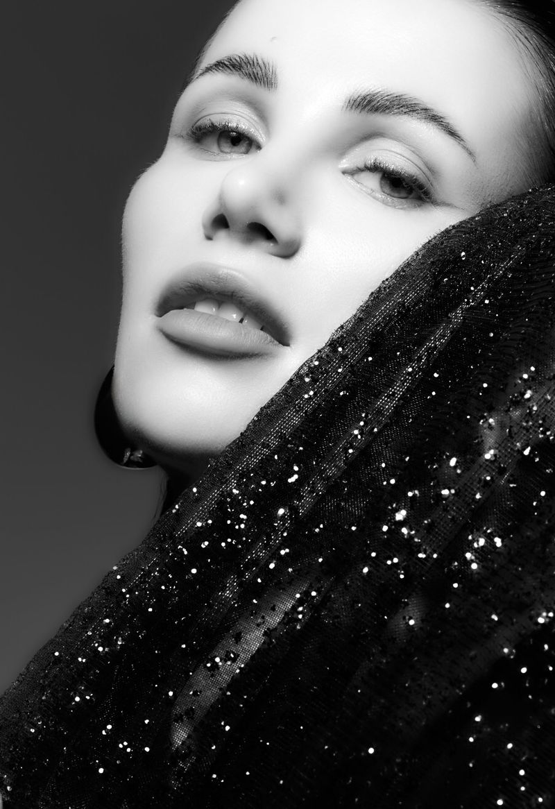 Black and white portrait of an adult woman with a sheer, sparkly black shawl draped on her face in a studio setting with diffused lighting.
