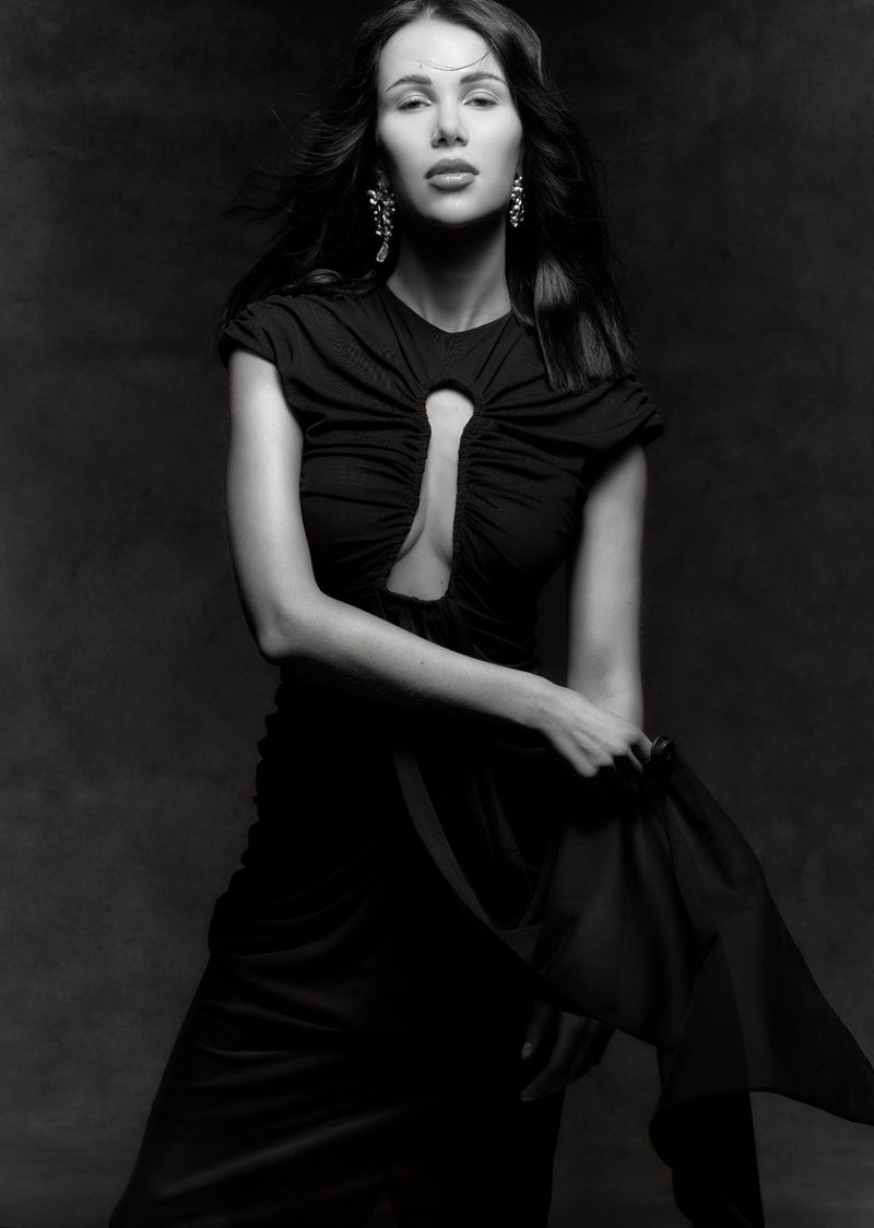 Elegant woman in a black dress poses against a dark background, showcasing fashion and portrait photography, with flowing fabric detail.