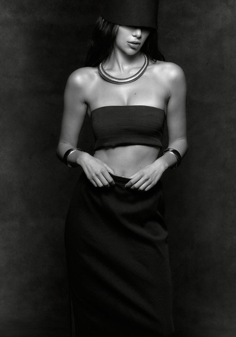 Monochrome portrait of a woman in a strapless top, skirt, and cylindrical hat, complemented by layered necklaces and bracelets against a dark backdrop.
