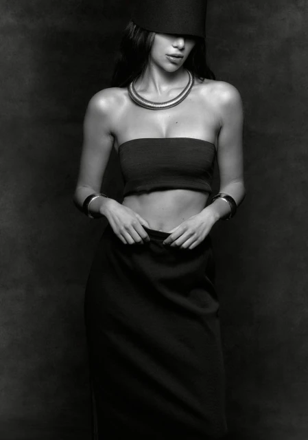 Monochrome portrait of a woman in a strapless top, skirt, and cylindrical hat, complemented by layered necklaces and bracelets against a dark backdrop.