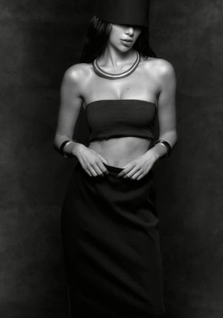 Monochrome portrait of a woman in a strapless top, skirt, and cylindrical hat, complemented by layered necklaces and bracelets against a dark backdrop.