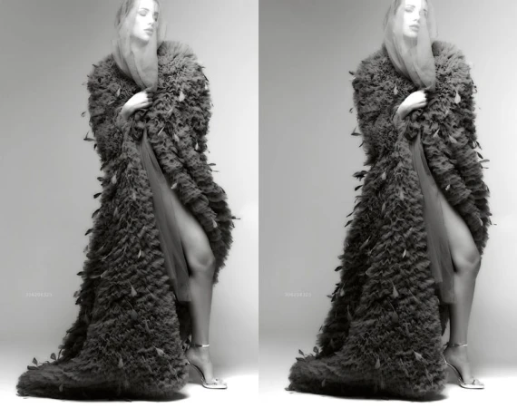 Monochrome image of a woman in a feathered coat with leg and heeled sandals visible, posing in a photo studio against a neutral backdrop.