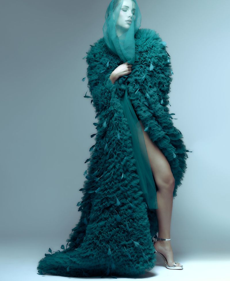 Model poses in a full-length feathered coat with sheer hood. Fashion photography with studio lighting and neutral background. 