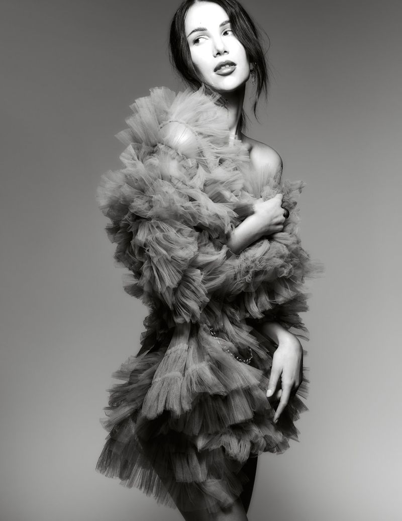 Model in a voluminous ruffled tulle dress posing against a gradient studio background in a black and white editorial portrait.