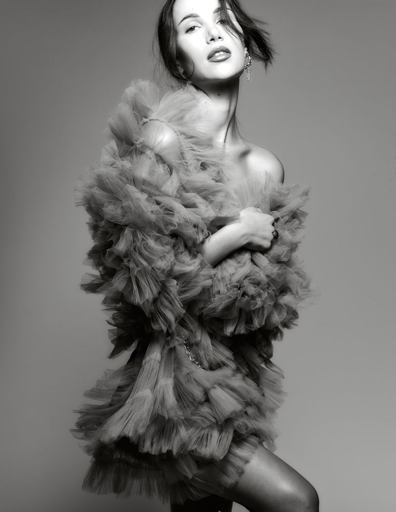Model Maria Gutjahr poses in a dramatic voluminous ruffled tulle mini dress against a neutral studio backdrop in black and white.