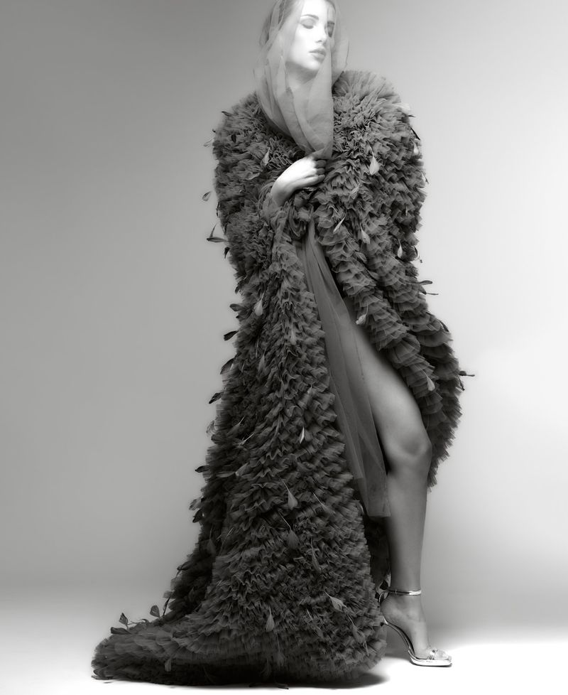 Monochrome fashion portrait of a woman in a feathered coat and sheer veil, posing in a studio with soft, diffused lighting.