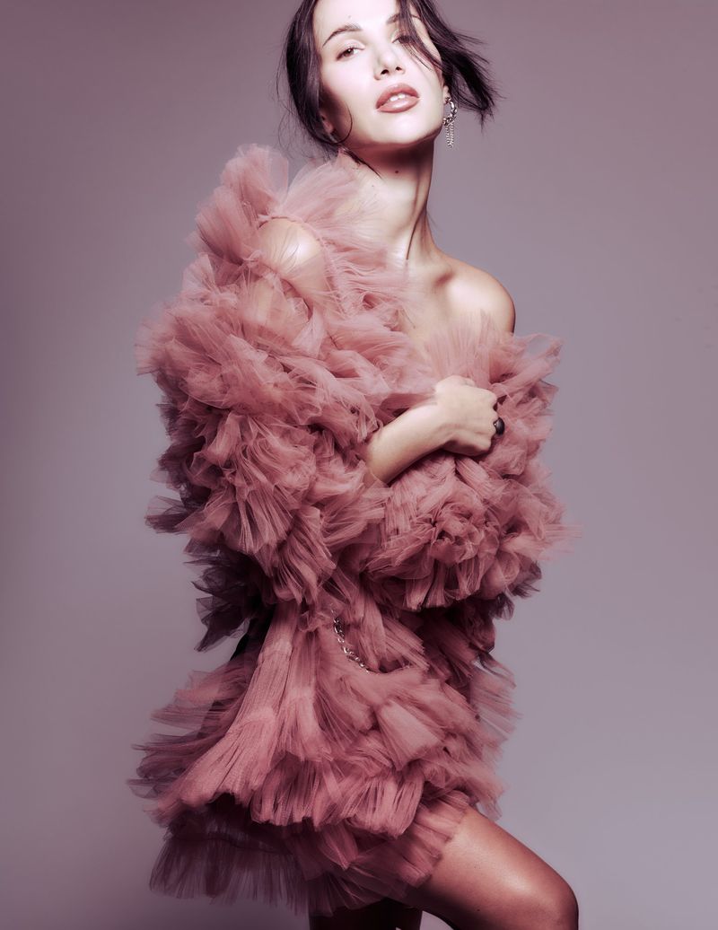 Elegant woman in a ruffled tulle dress strikes a pose against a grey background, showcasing a fashionable and textured look with soft lighting.