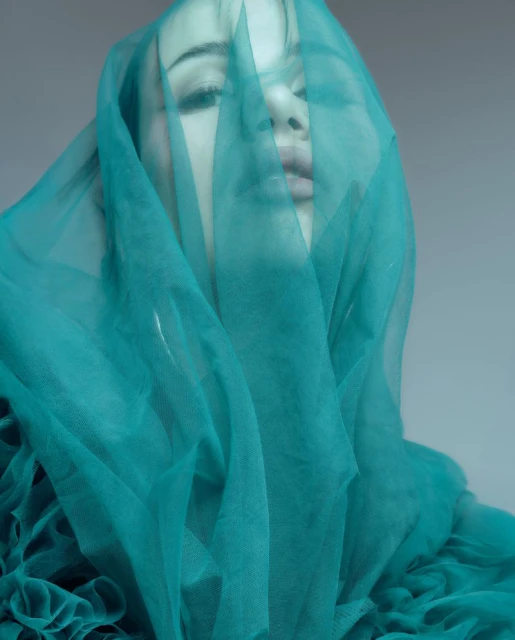 Close-up studio shot of a woman partially veiled with a teal chiffon fabric, emphasizing ethereal beauty, and soft lighting.