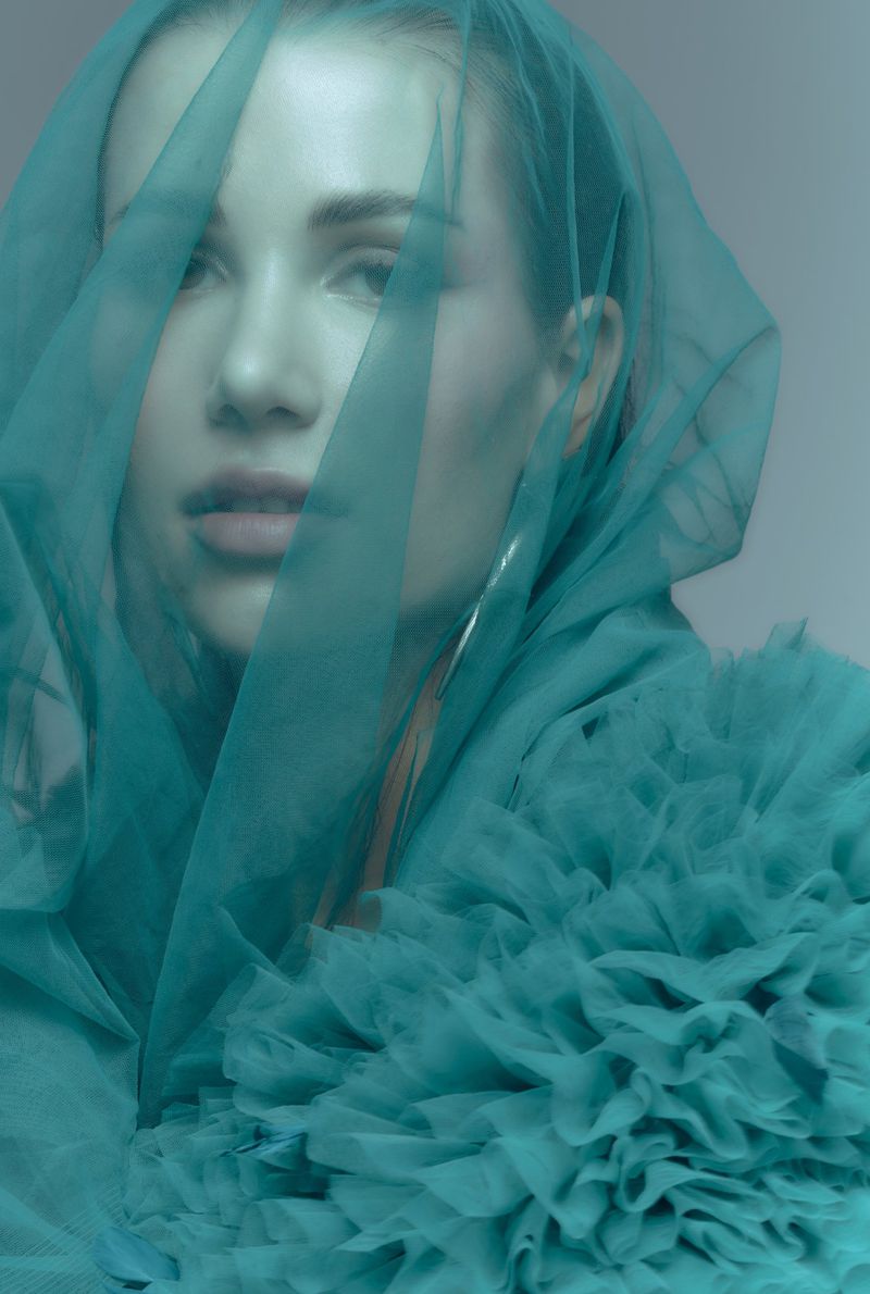Close-up portrait of a young woman with light skin and teal fabric draped over her head and shoulders in a studio setting.