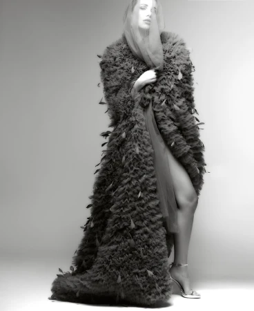 Model Maria Gutjahr posing in a dramatic floor-length feathered ruffle coat and sheer headpiece against a studio backdrop.