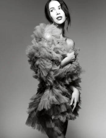 Model in a voluminous ruffled tulle dress posing against a gradient studio background in a black and white editorial portrait.