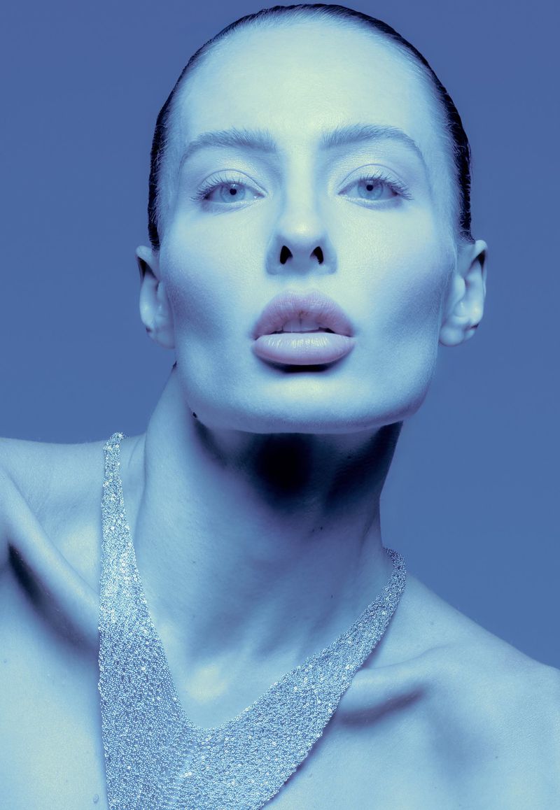 Close-up studio shot of a fair-skinned adult woman with pursed lips wearing a mesh top against a gradient blue background.