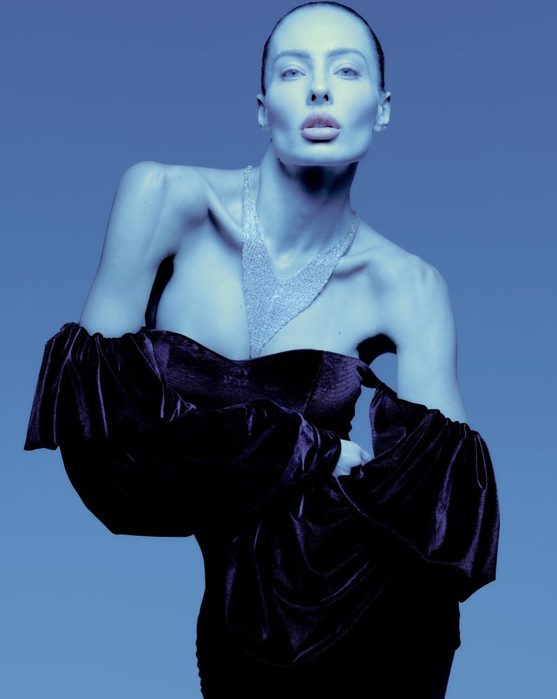 Model in off-the-shoulder velvet dress wearing a sparkling necklace against a blue studio background