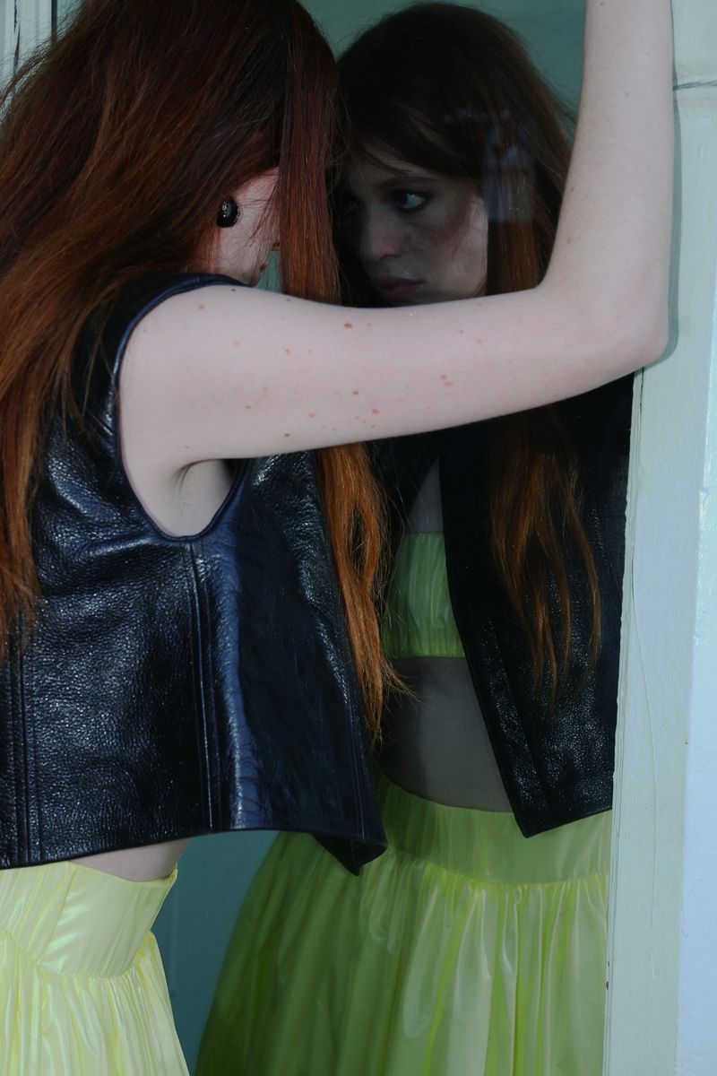 A red-haired woman in a black vest and yellow skirt stands next to a mirror, reflecting her face and outfit in an indoor setting.