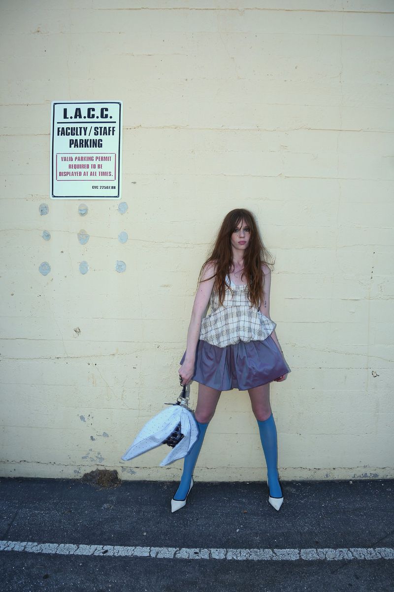 A long-haired woman stands in front of a pale wall, wearing a short purple skirt, blue socks, white heels, and holding an umbrella.
