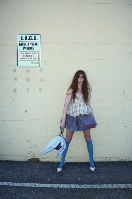A long-haired woman stands in front of a pale wall, wearing a short purple skirt, blue socks, white heels, and holding an umbrella.