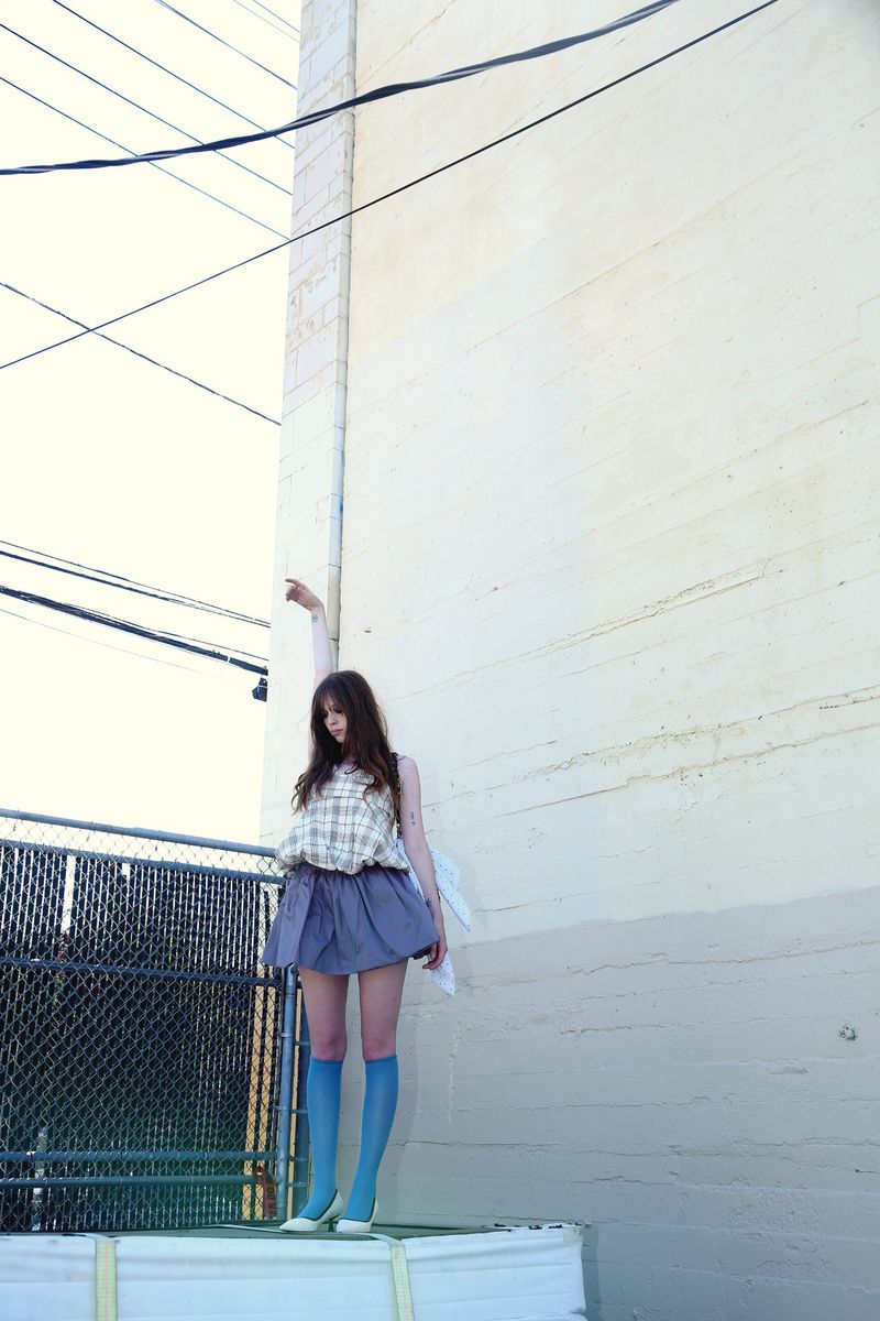A young adult woman in a blouse, skirt, knee-high socks, and flats poses on a white platform against an urban concrete wall backdrop.