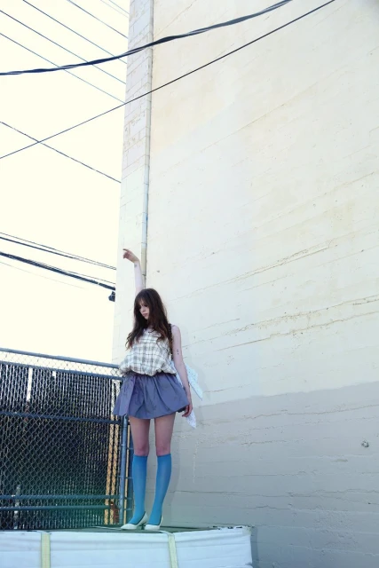 A young adult woman in a blouse, skirt, knee-high socks, and flats poses on a white platform against an urban concrete wall backdrop.