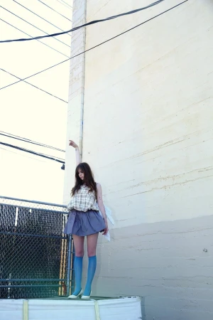 A young adult woman in a blouse, skirt, knee-high socks, and flats poses on a white platform against an urban concrete wall backdrop.