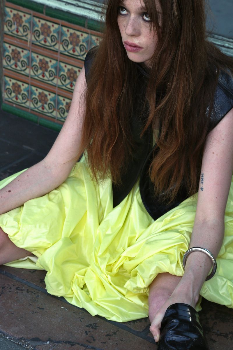 A woman with long red hair is seated on a patterned floor, wearing a black vest and a voluminous, bright yellow dress.