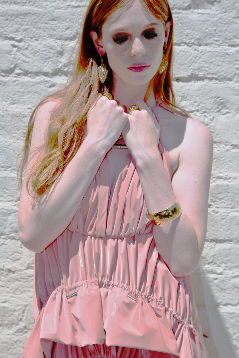 Portrait of a woman with long red hair wearing a light pink dress and gold jewelry standing against a white brick wall in daylight.
