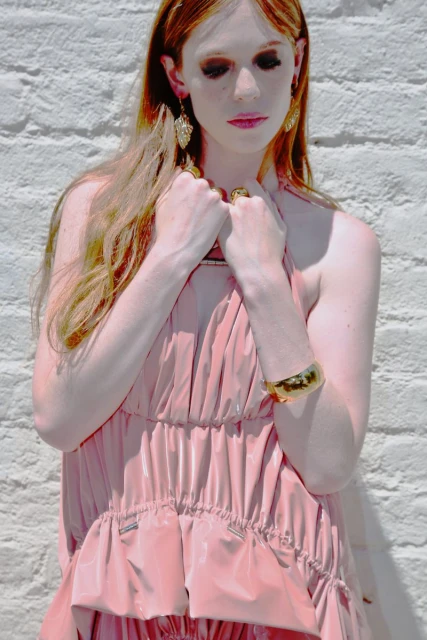 Portrait of a woman with long red hair wearing a light pink dress and gold jewelry standing against a white brick wall in daylight.
