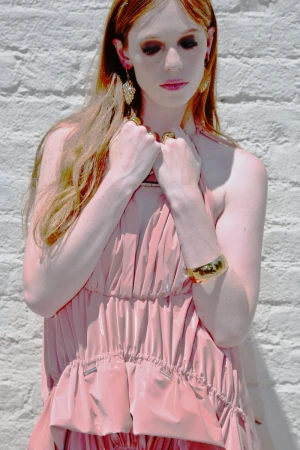 Portrait of a woman with long red hair wearing a light pink dress and gold jewelry standing against a white brick wall in daylight.