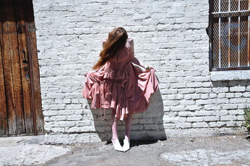 Woman with long red hair stands in pink dress against a white brick wall next to a wooden door, wearing white heels in bright sunlight.