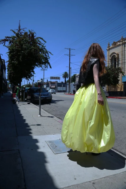 An adult female in a yellow gown and black top walking down a city street on a sunny day, captured in a medium shot with a blue sky.