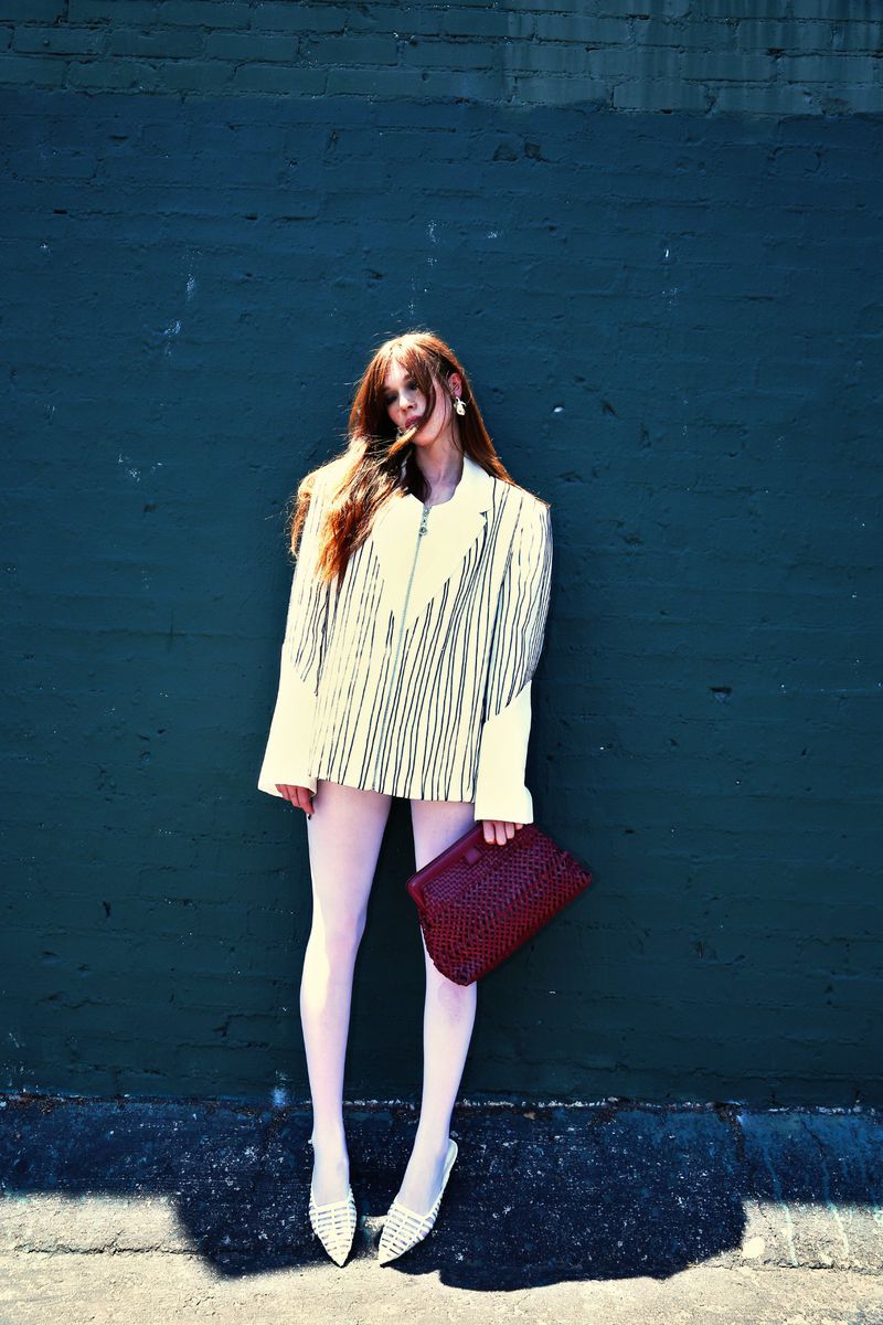 A stylish woman poses against a dark blue wall, wearing a striped blazer, light pink tights, and woven flats, holding a dark red woven clutch.